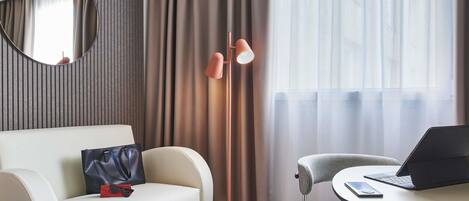Een moderne hotelkamer met een sofa, een klein rond tafeltje met een laptop, een stoel, een lamp en een spiegel.
