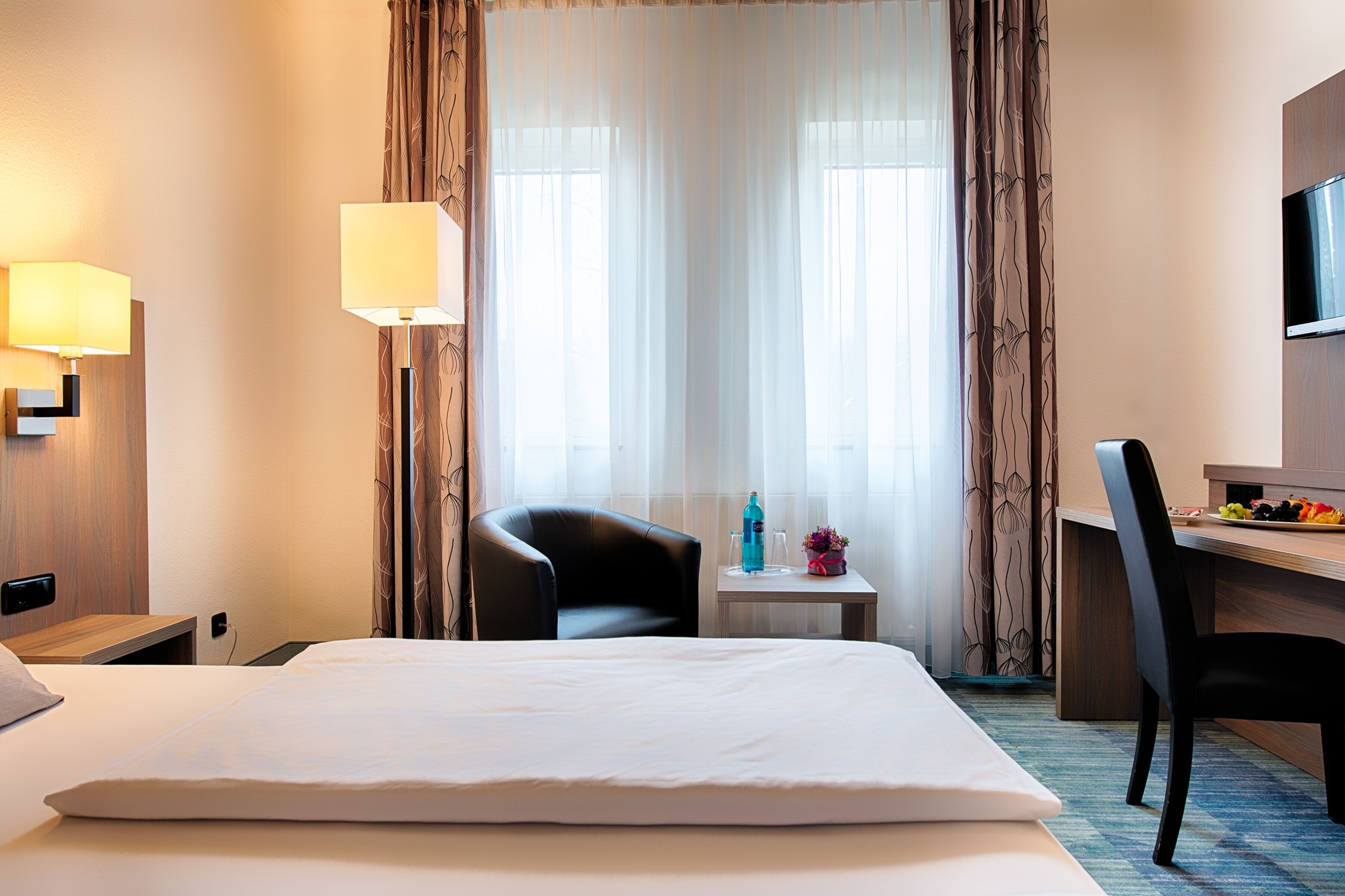 A hotel room with a bed, a desk, a chair, a TV, and a window with curtains.