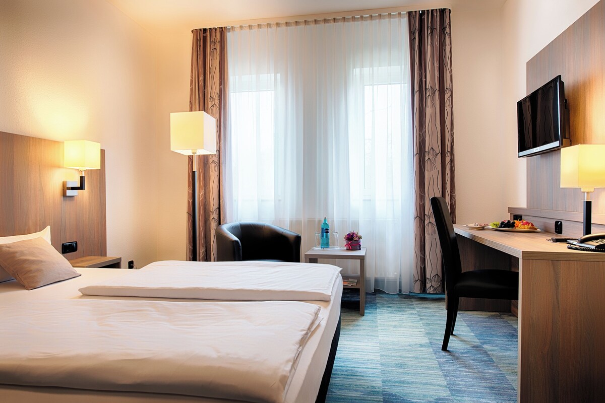A hotel room with a large bed, a desk with a chair, a TV mounted on the wall, and a window with curtains.