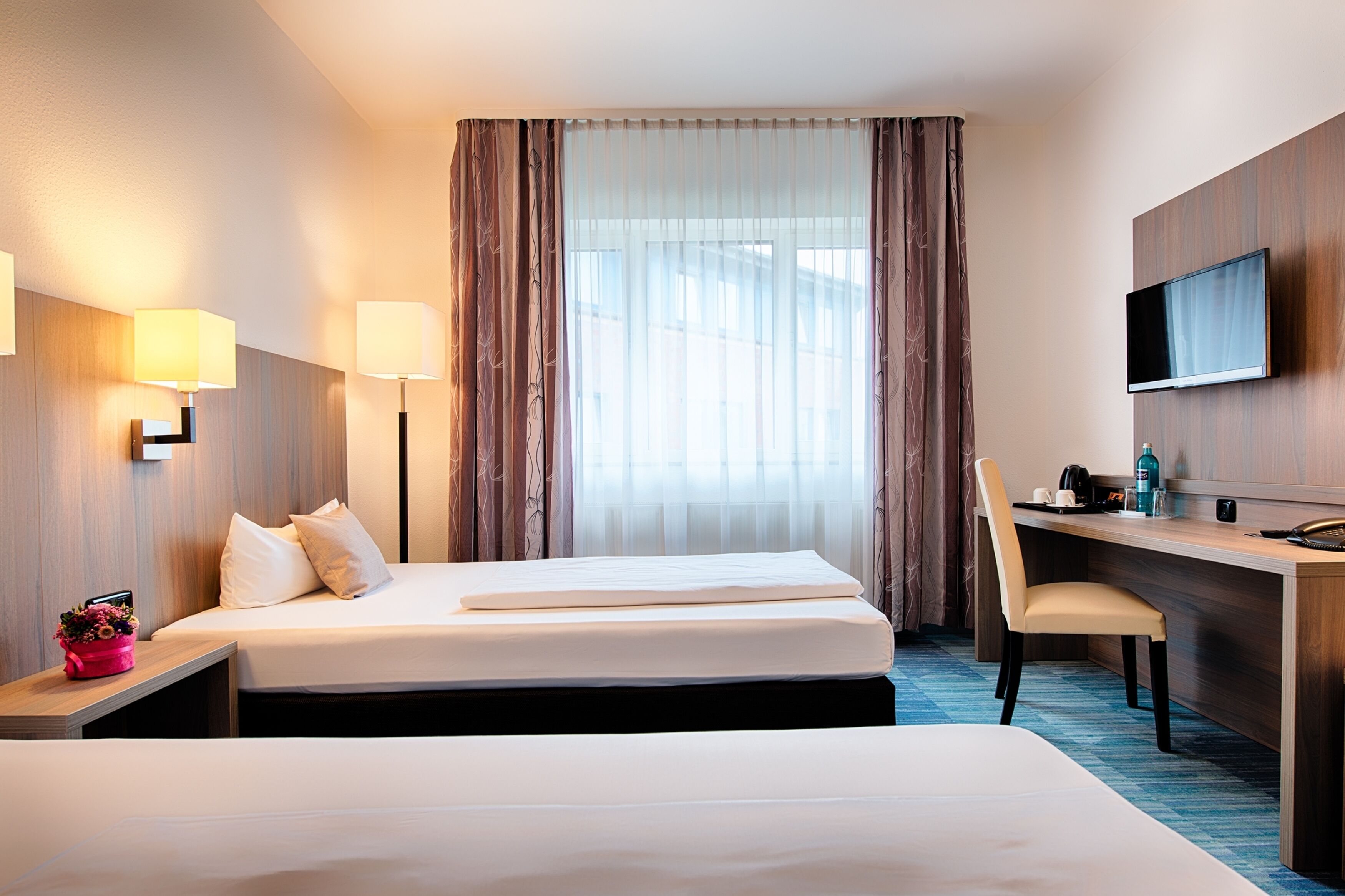Ein Hotelzimmer mit zwei Betten, einem Schreibtisch, einem Sessel, einem Fernseher und einem Vorhangfenster.