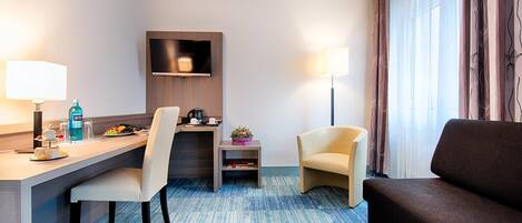 A hotel room with a desk, two chairs, a sofa, a flat-screen TV, and a small table with a lamp.