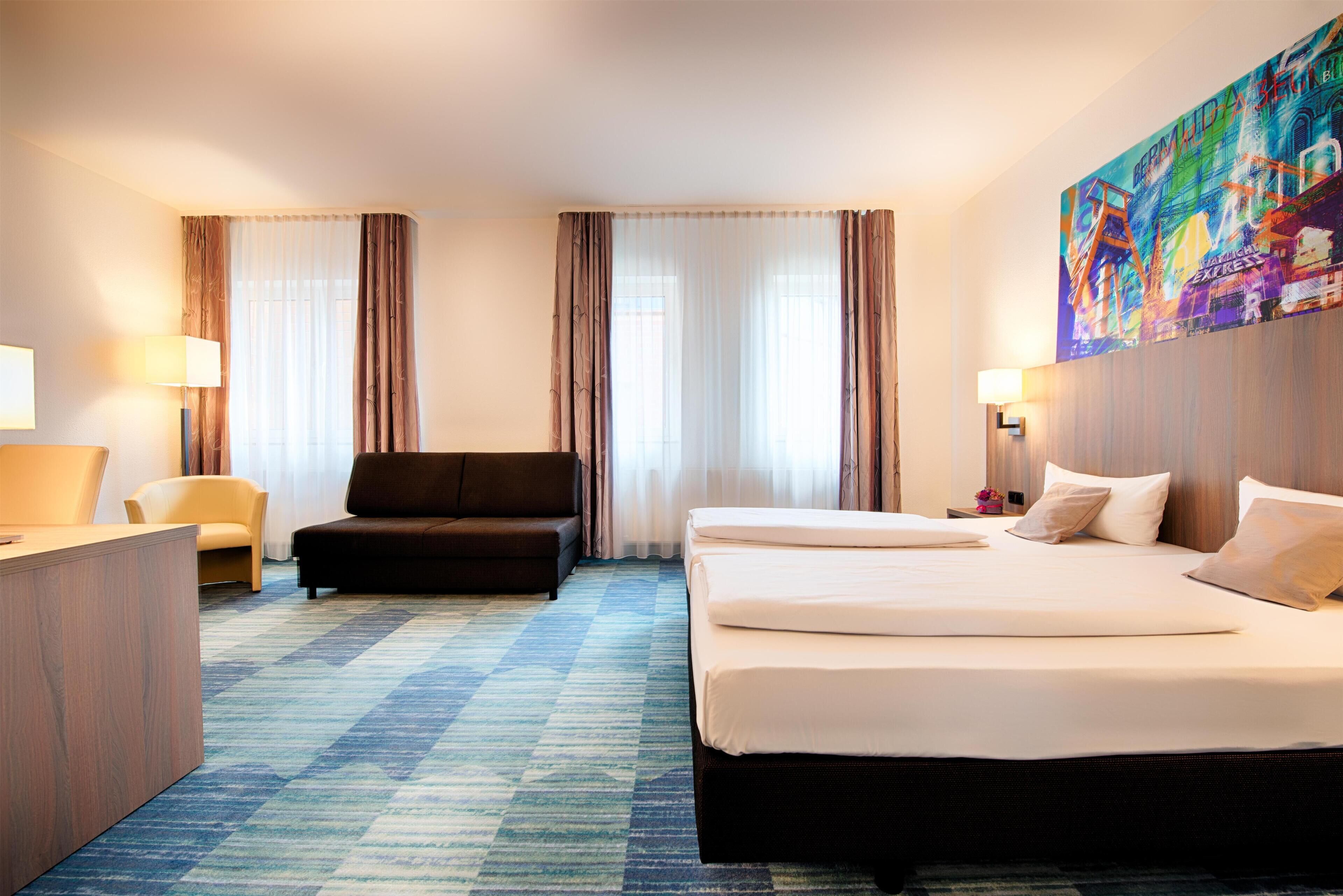 A hotel room with a large bed, a desk, a chair, and a couch. There are curtains on the windows and a painting on the wall.