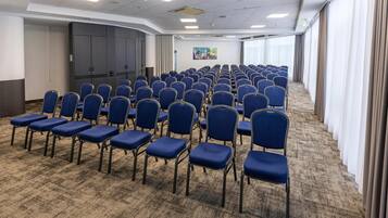 A conference room with rows of blue chairs, a large grey cabinet, and a colorful painting on the wall.