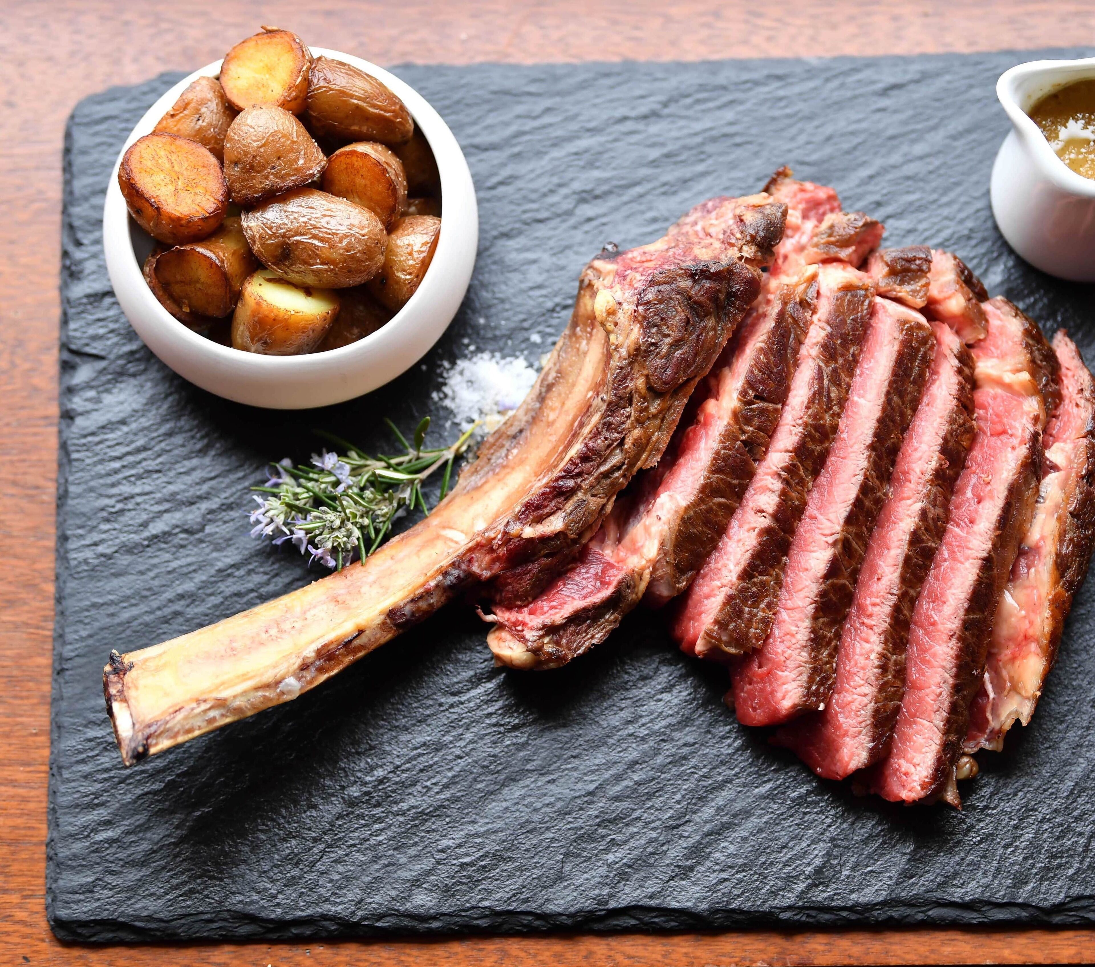 A steak with a bone, sliced, and a bowl of roasted potatoes on a slate board.