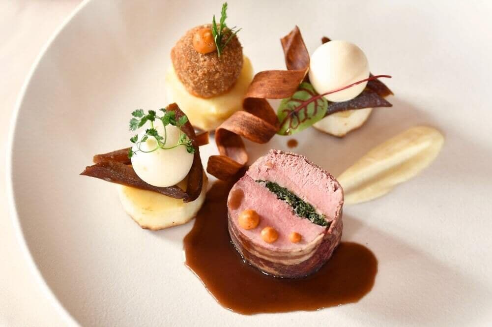 Een bord met verschillende elementen, waaronder een rond vleesgerecht, een bolvormig element en een cilindervormig stuk, gegarneerd met kruiden en saus.