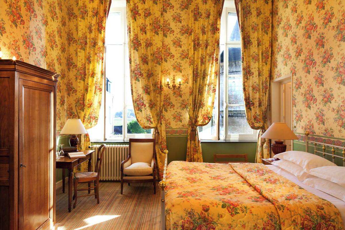 A room with floral wallpaper, a bed with a floral bedspread, a chair, a desk with a lamp, and a radiator.