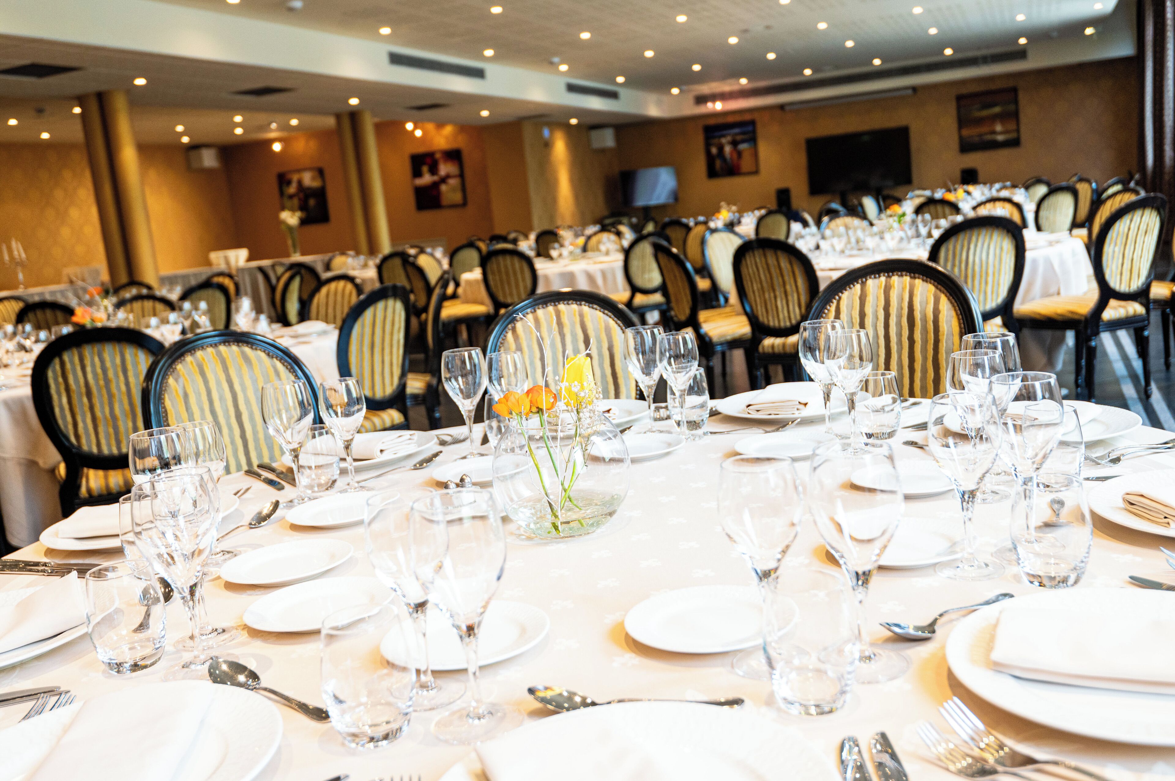 A banquet hall with multiple round tables set for dining, each with white tablecloths, silverware, and clear glassware.