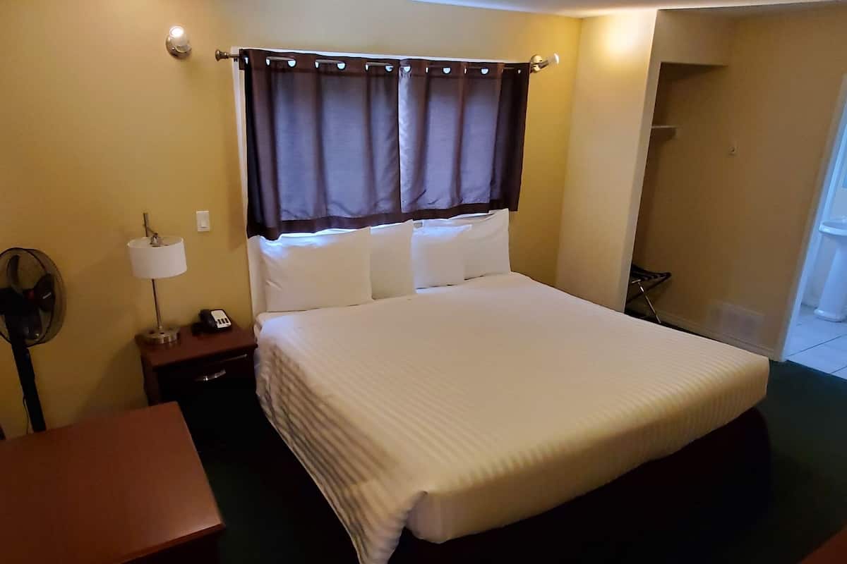 A hotel room with a bed, a bedside table with a lamp, a fan, and a bathroom visible in the background.