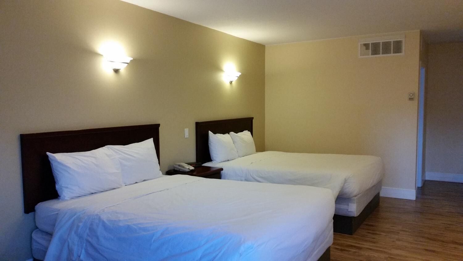 A hotel room with two beds, each with a wooden headboard and white bedding. There are wall-mounted light fixtures and a visible vent on the wall.