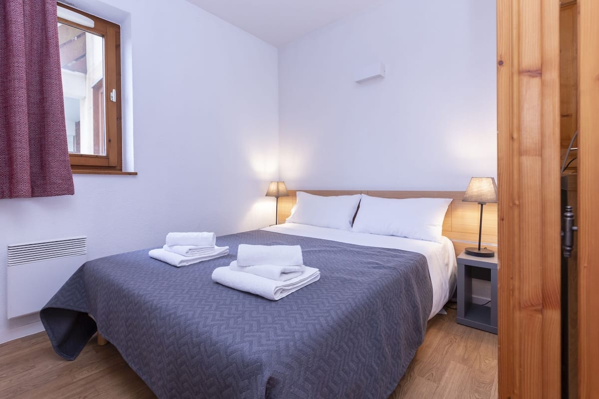 A neatly made bed with a grey patterned bedspread, two white towels, and two bedside lamps.