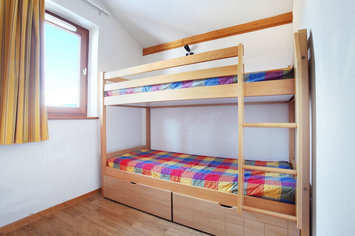 A bunk bed with a colorful quilt, wooden drawers, and a window with curtains.
