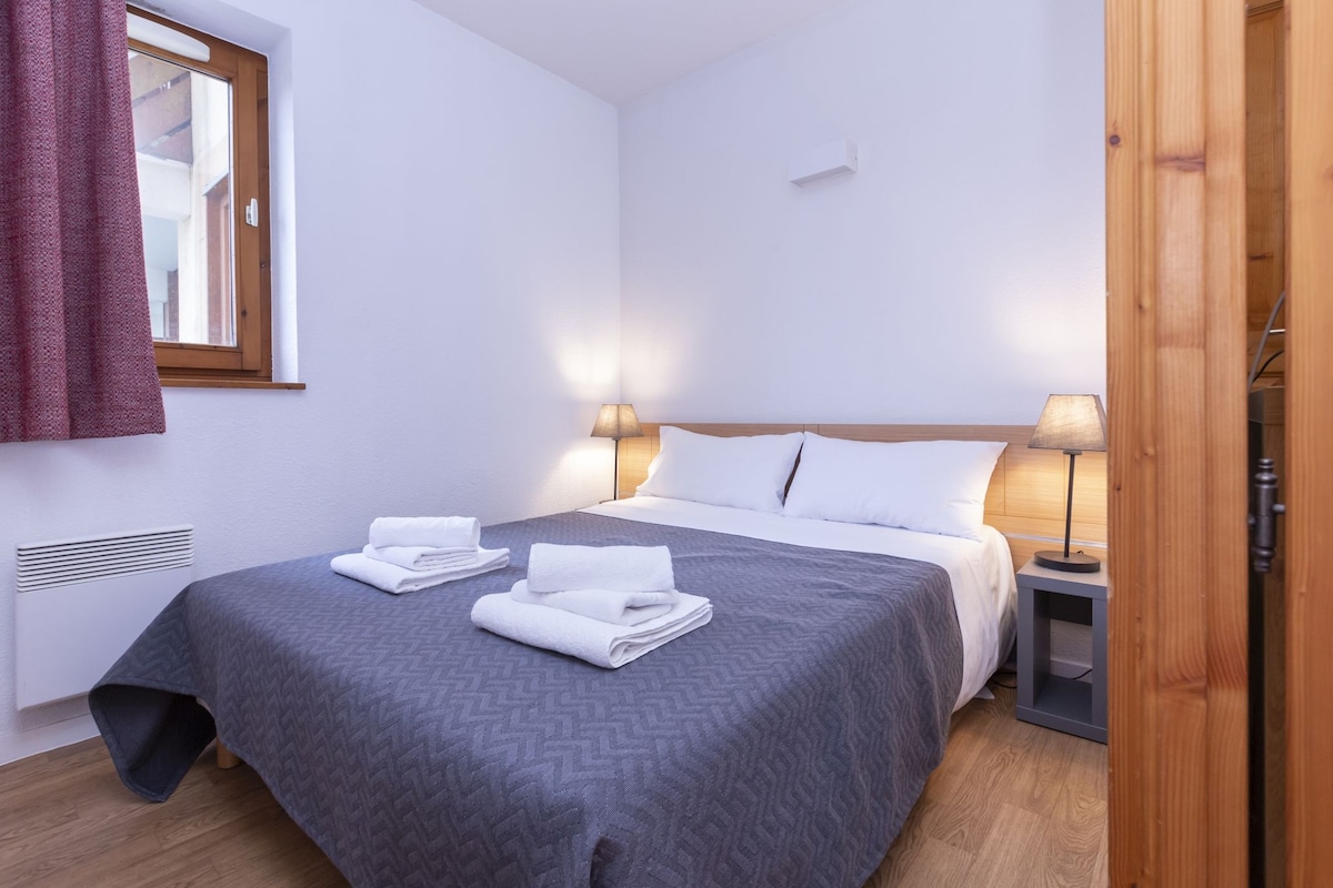 A neatly made bed with a grey patterned bedspread, two white towels, and two bedside lamps.
