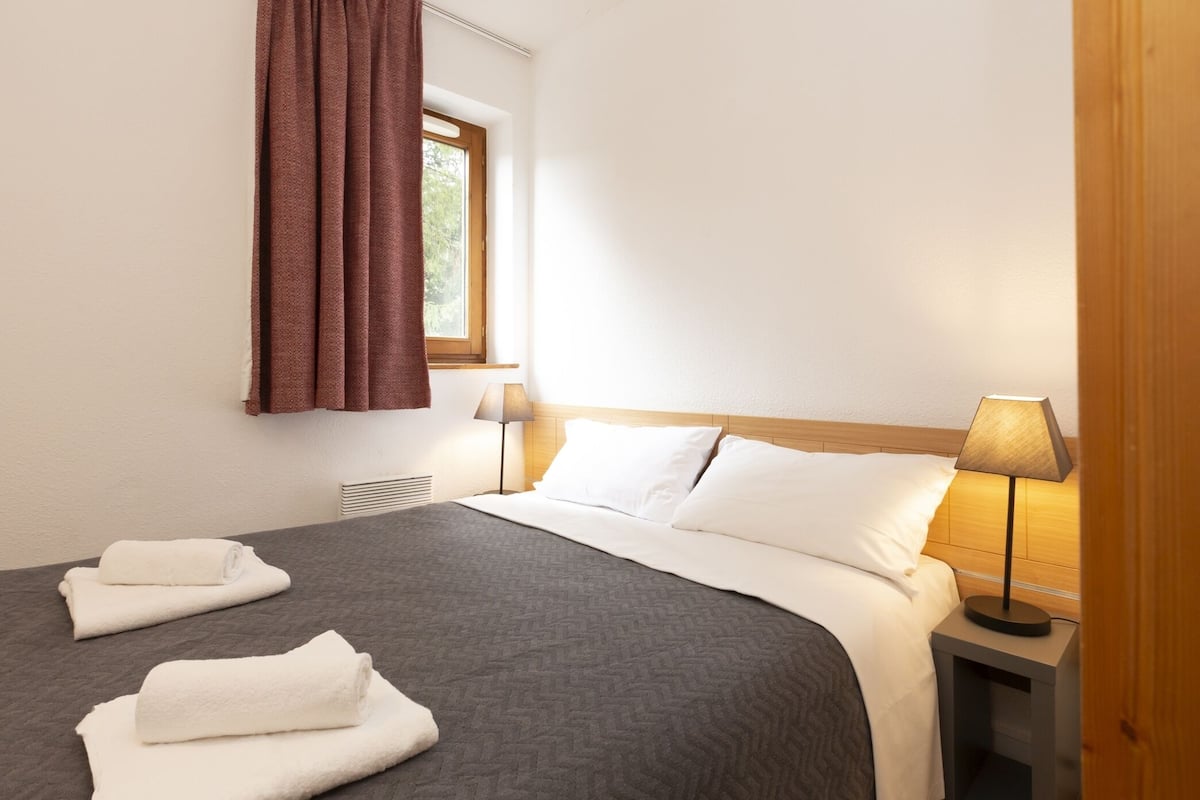 A hotel room with a bed, two pillows, a nightstand with a lamp, and a window with curtains.