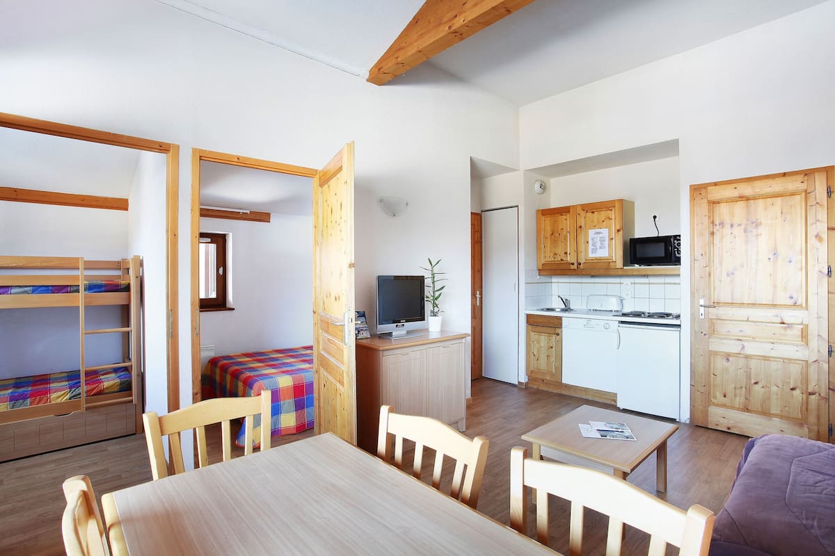 A compact room with bunk beds, a dining table, wooden chairs, a small kitchen area with a microwave, and a cabinet.