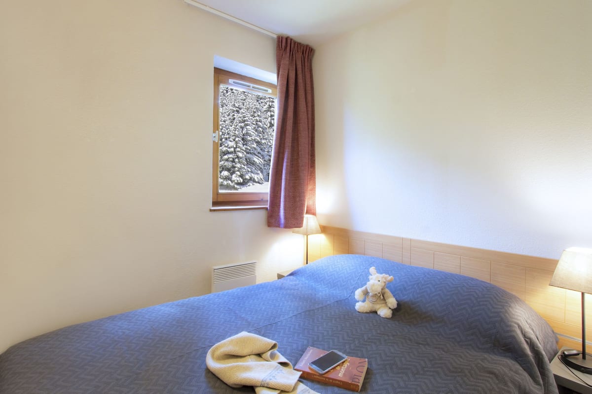 A hotel room with a bed, a window with a view of trees, a lamp, a stuffed toy, and a book.