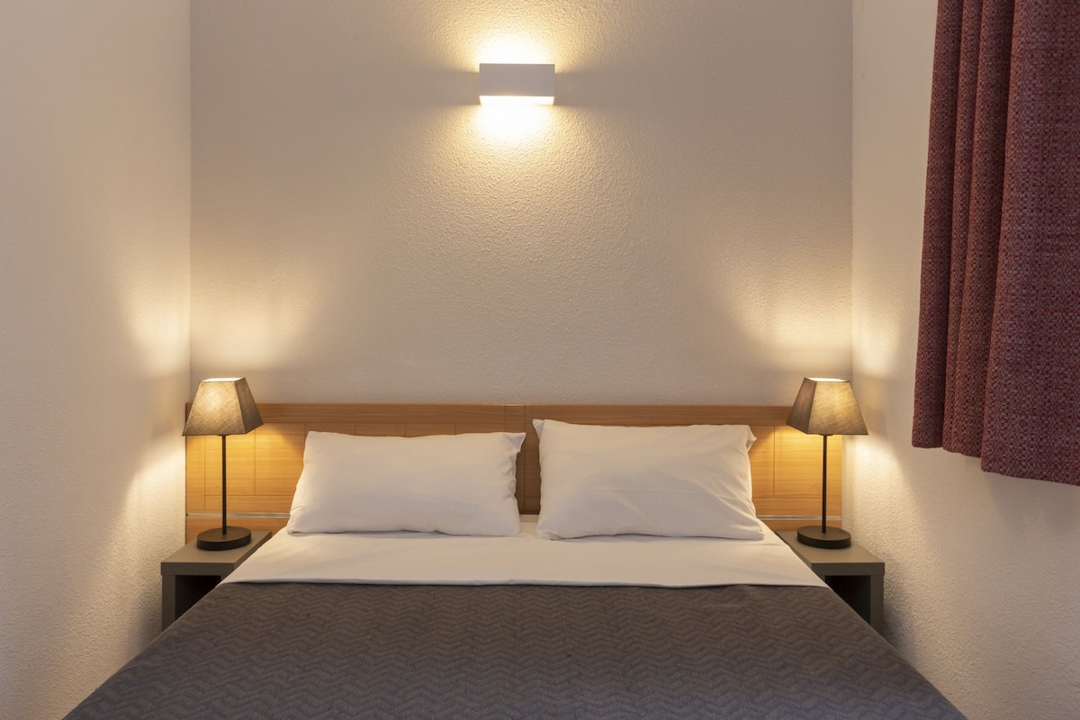 A hotel room with a bed, two bedside lamps, a wall-mounted light, and a window with curtains.
