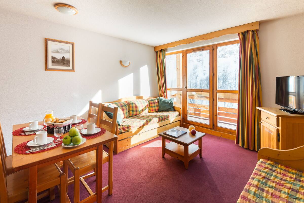 Une pièce chaleureuse avec une table à manger en bois, mise pour le petit-déjeuner, un canapé agrémenté de coussins colorés, une petite table en bois avec un livre, et un téléviseur posé sur une commode en bois.