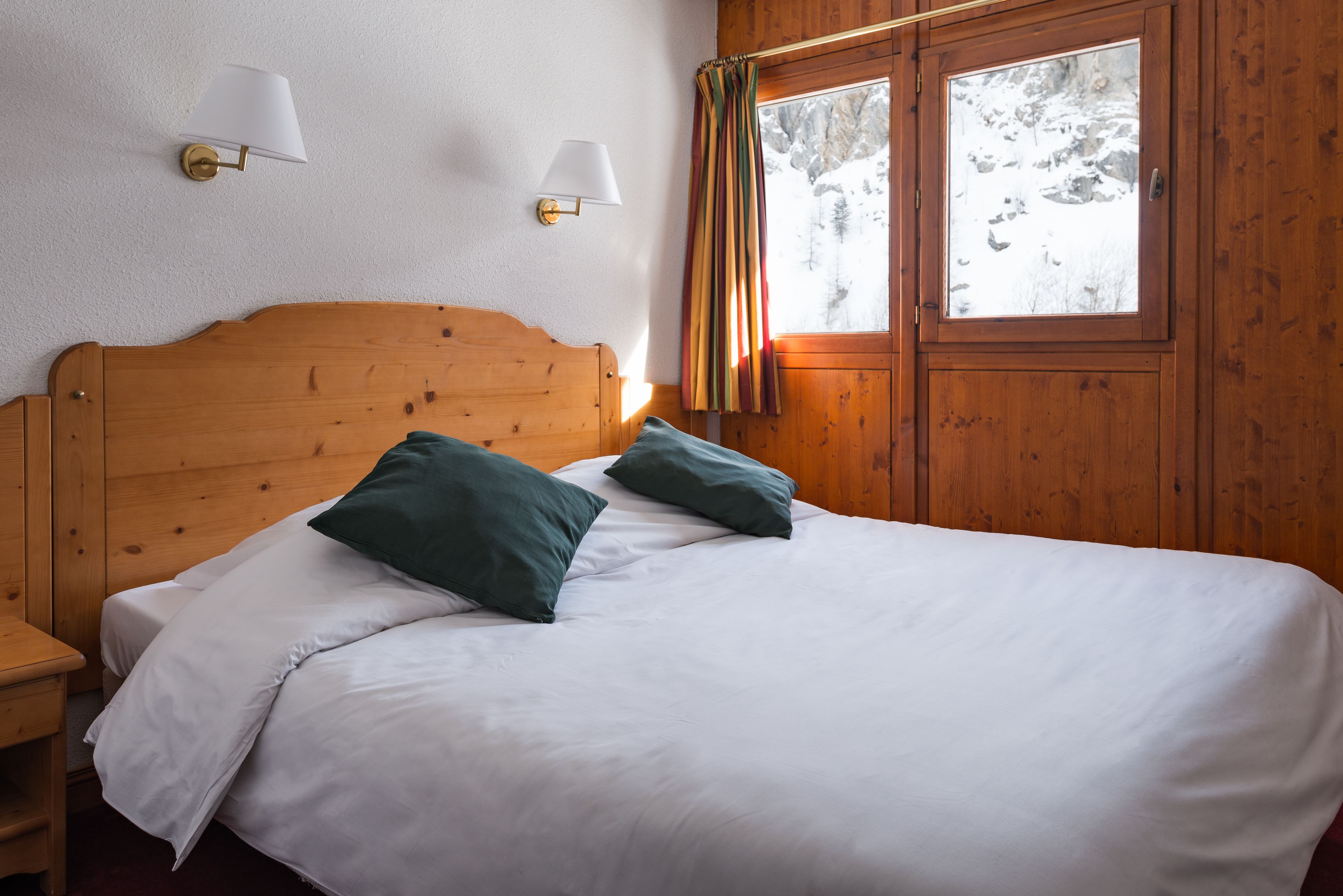 Un lit en bois avec une literie blanche et deux oreillers, une table de chevet et une fenêtre avec des rideaux.