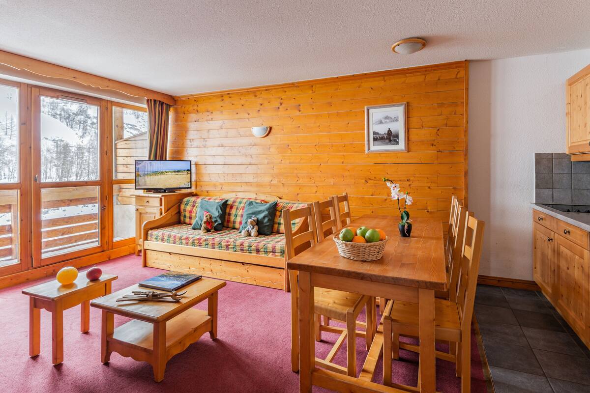 L’intérieur chaleureux d’un chalet avec une table à manger en bois, un canapé agrémenté de coussins colorés, un téléviseur posé sur un meuble en bois et un coin cuisine doté de placards en bois.
