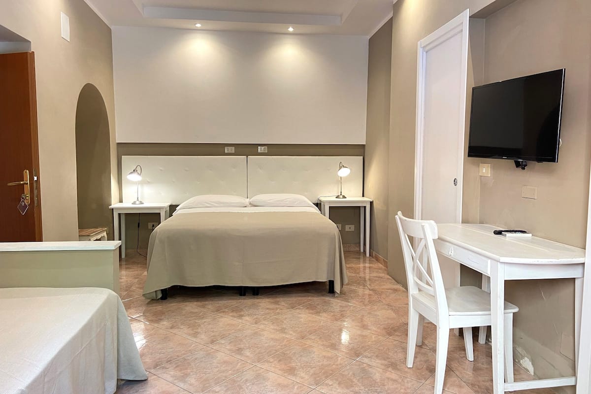 A hotel room with a bed, bedside tables, a desk with a chair, a television, and a door.