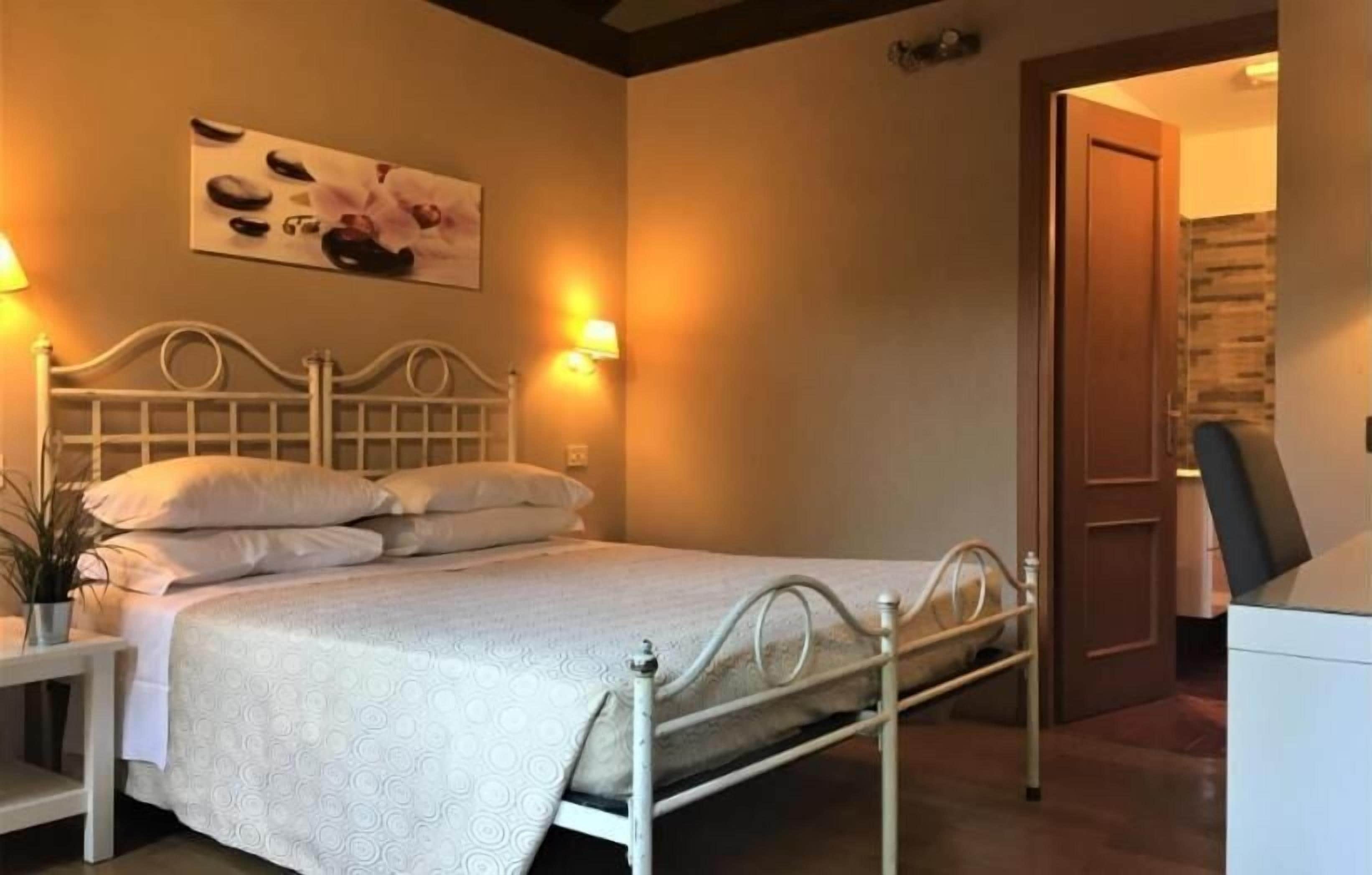 Una cama bien tendida con cabecera y piecera blancos, una mesita de noche con una lámpara, y una obra de arte colgada en la pared sobre la cama.