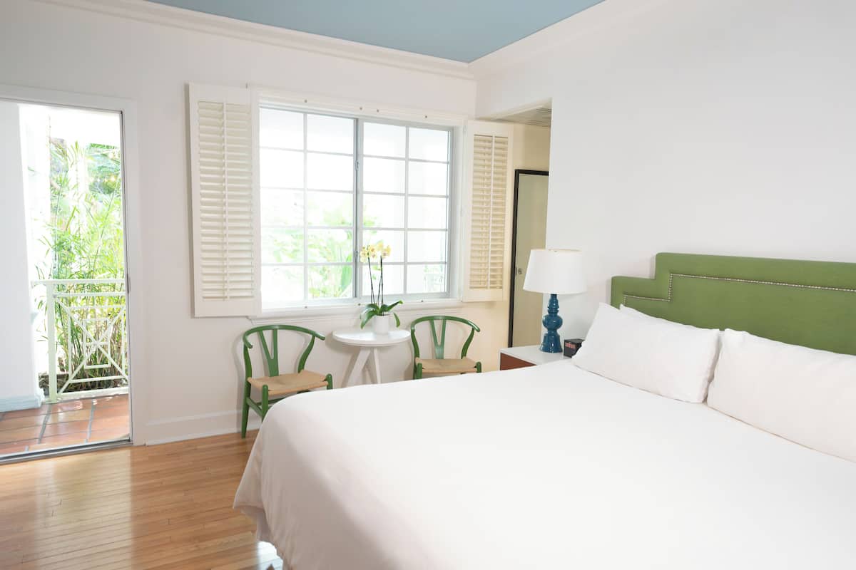A hotel room with a bed, a small table with chairs, a window with shutters, and a view of greenery outside.