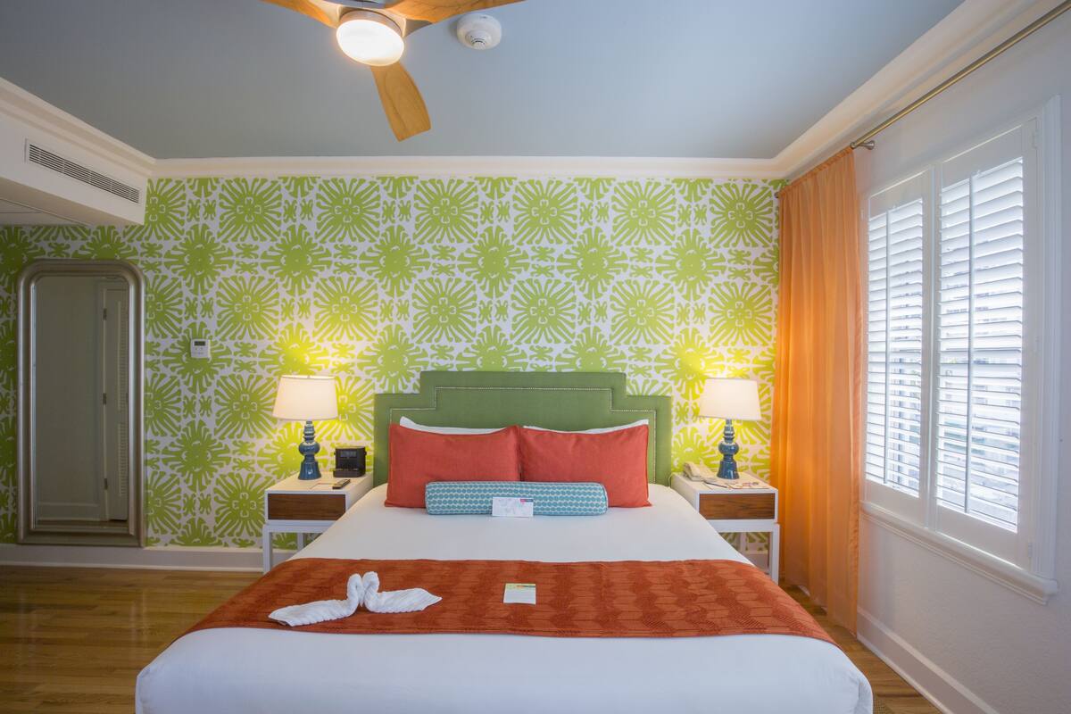 A hotel room with a green patterned wall, a bed with red and blue pillows, two bedside lamps, a wooden ceiling fan, and a window with blinds.