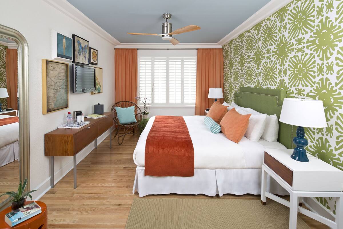 A hotel room with a bed, bedside tables, a desk, a chair, a ceiling fan, and a window with curtains.