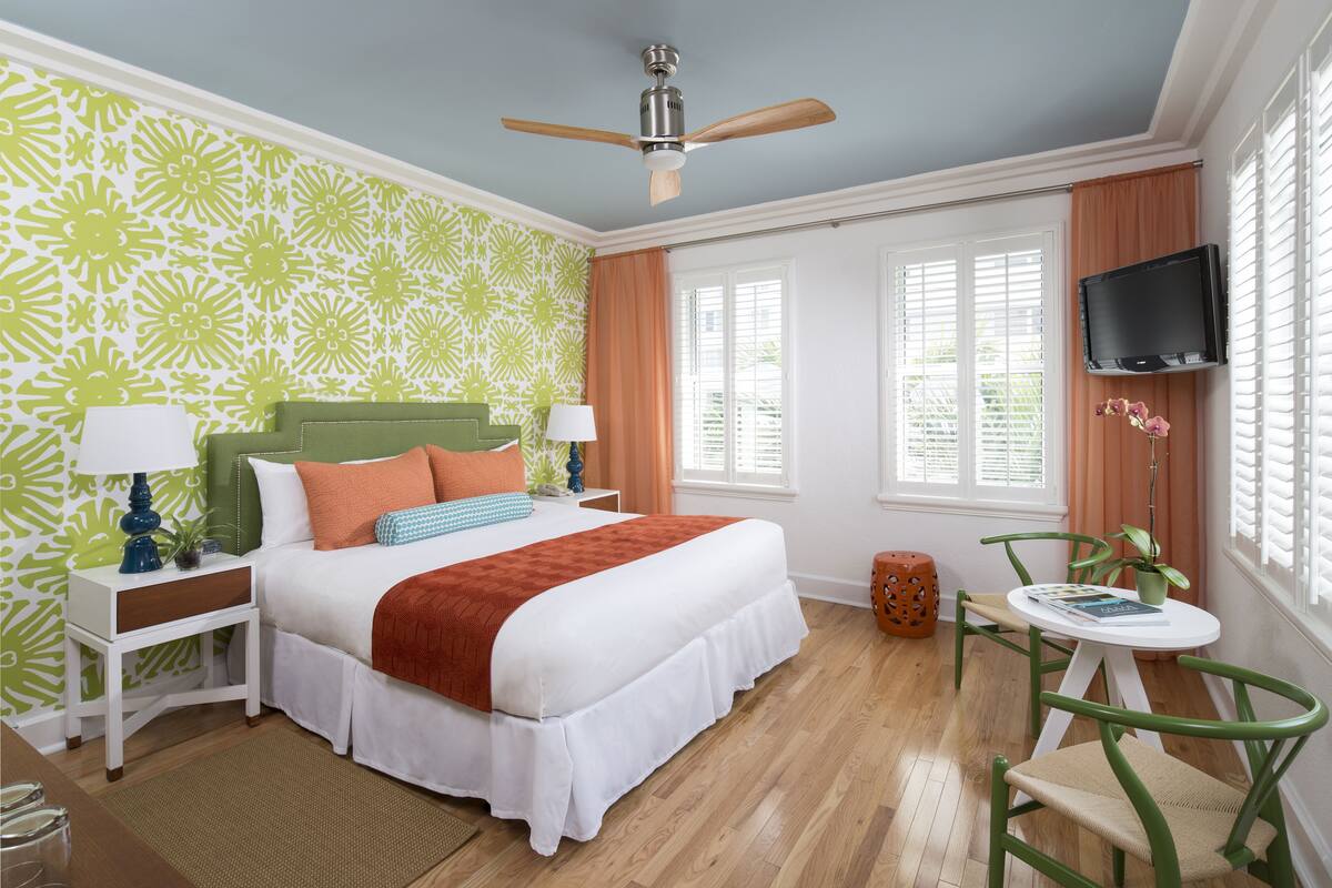 A hotel room with a bed, a ceiling fan, a TV mounted on the wall, and a window with blinds.