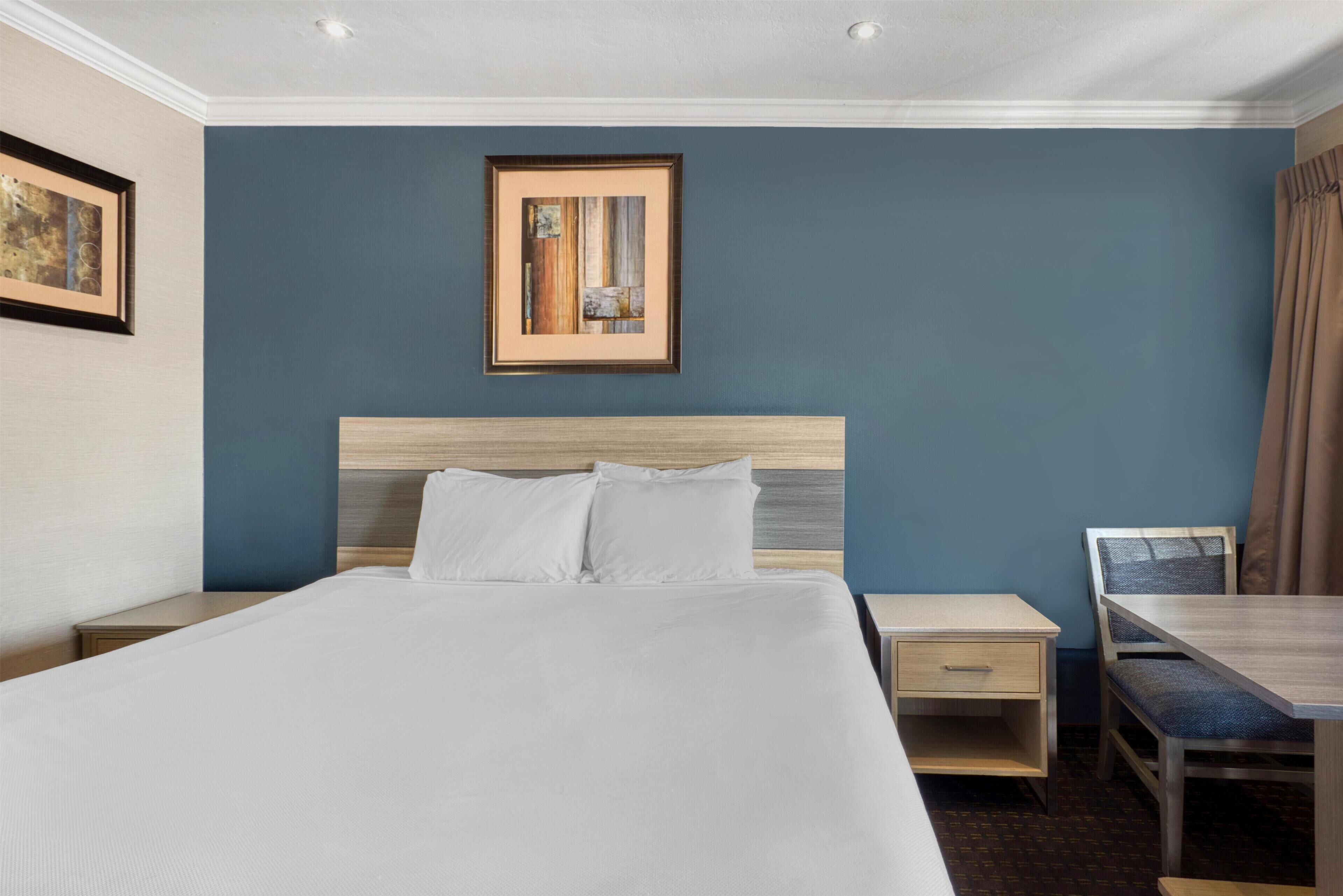 A hotel room with a large bed, a wooden headboard, a nightstand, a desk, and a chair. There is a framed picture on the wall above the bed.