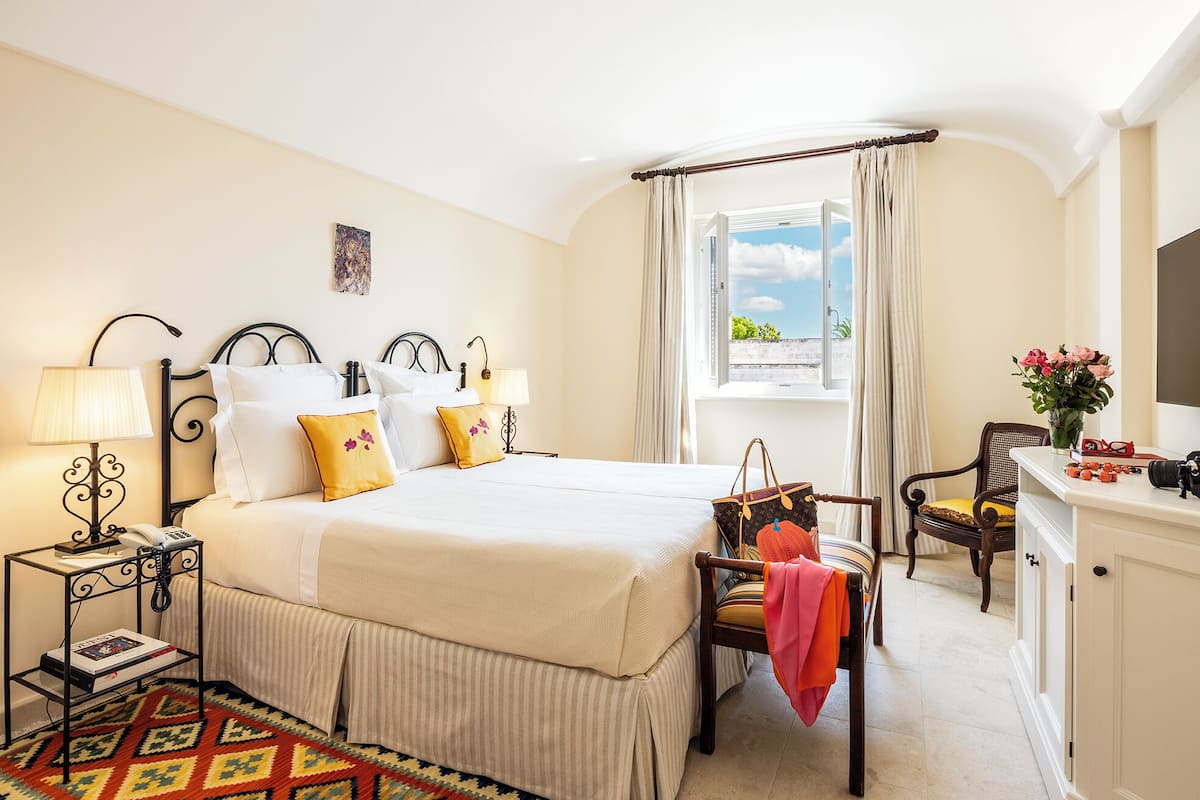 Habitación de hotel con cama, mesita de noche, lámpara, silla, escritorio y ventana con cortinas.