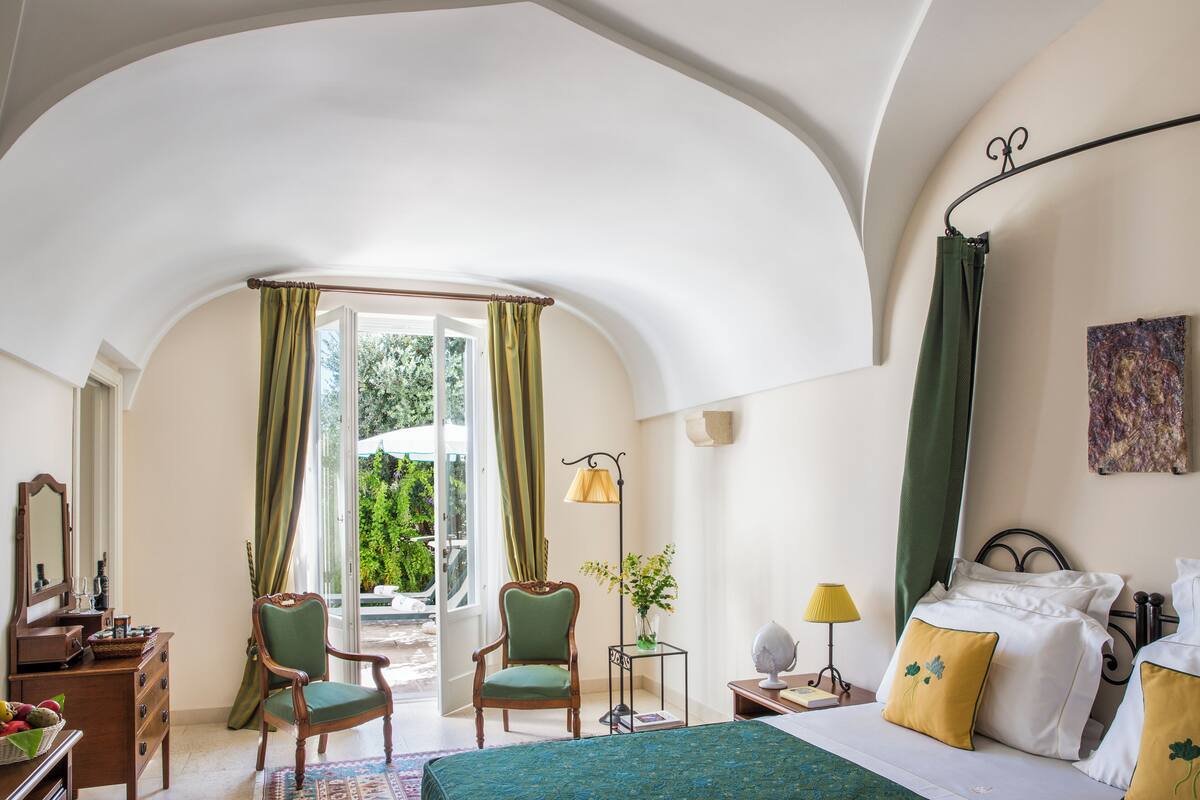 Un dormitorio con cama, dos sillones, un escritorio, una lámpara y vistas al verdor exterior.