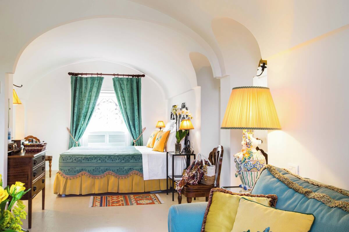 Una habitación con cama, sofá, lámpara, una mesita y un jarrón decorativo.