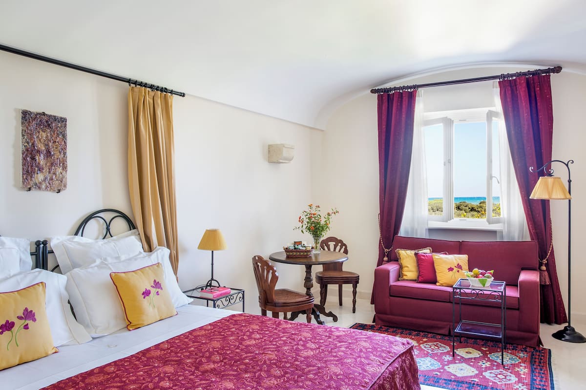 Una habitación de hotel con cama, sofá, mesa de comedor con sillas, ventana con cortinas y una lámpara.