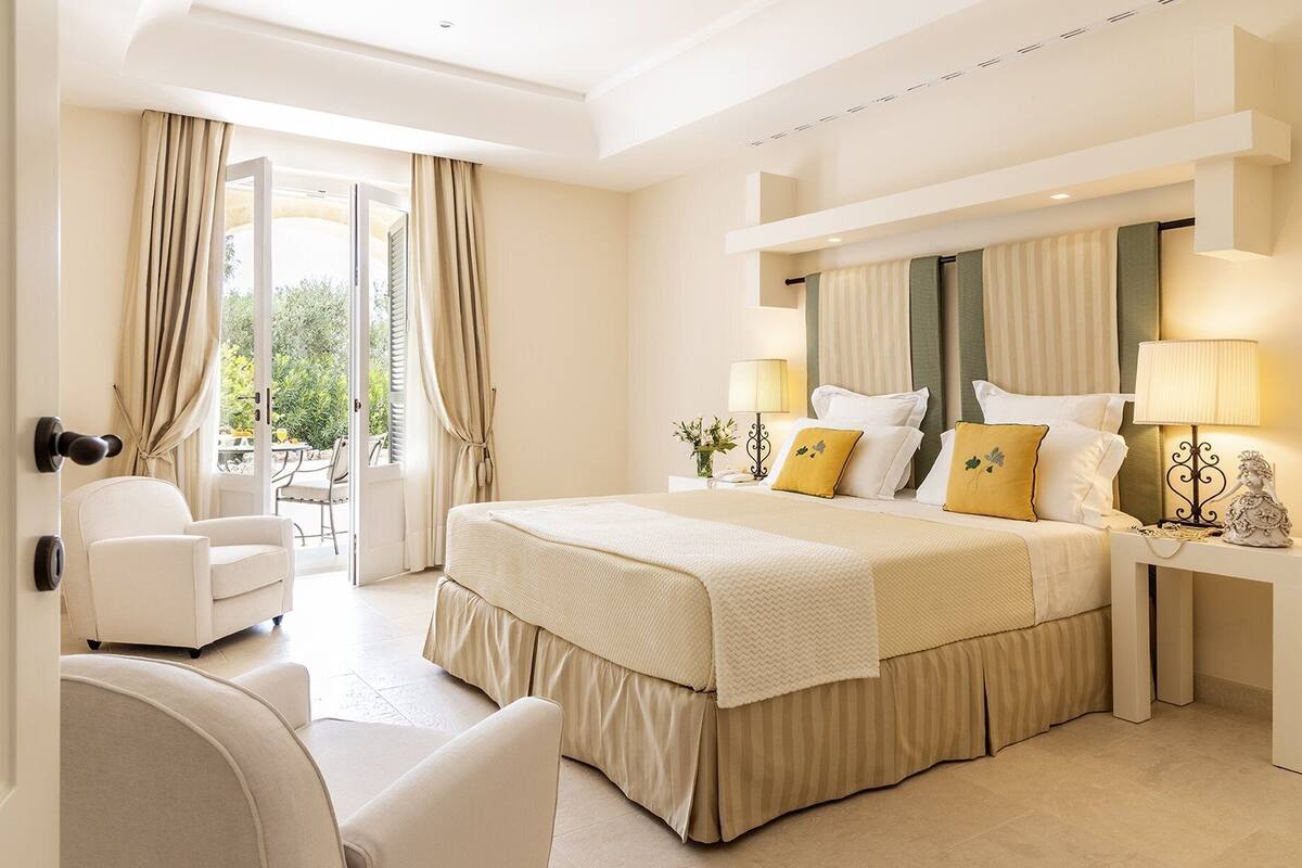 Habitación de hotel con una cama grande, zona de estar con una silla, escritorio con lámpara y vistas a la vegetación exterior.
