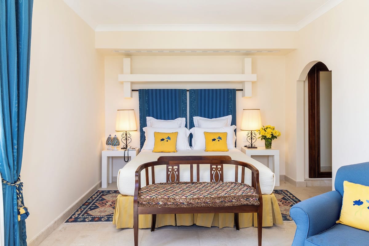 Habitación de hotel con cama, dos lámparas de noche, una silla, una mesita y una ventana con cortinas azules.