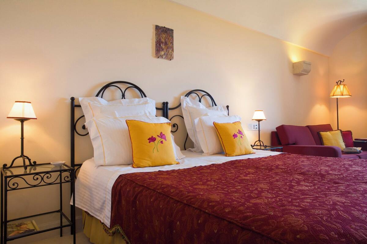 Una cama doble con sábanas blancas y almohadas de flores amarillas, una colcha estampada roja y dos mesitas de noche con lámparas.
