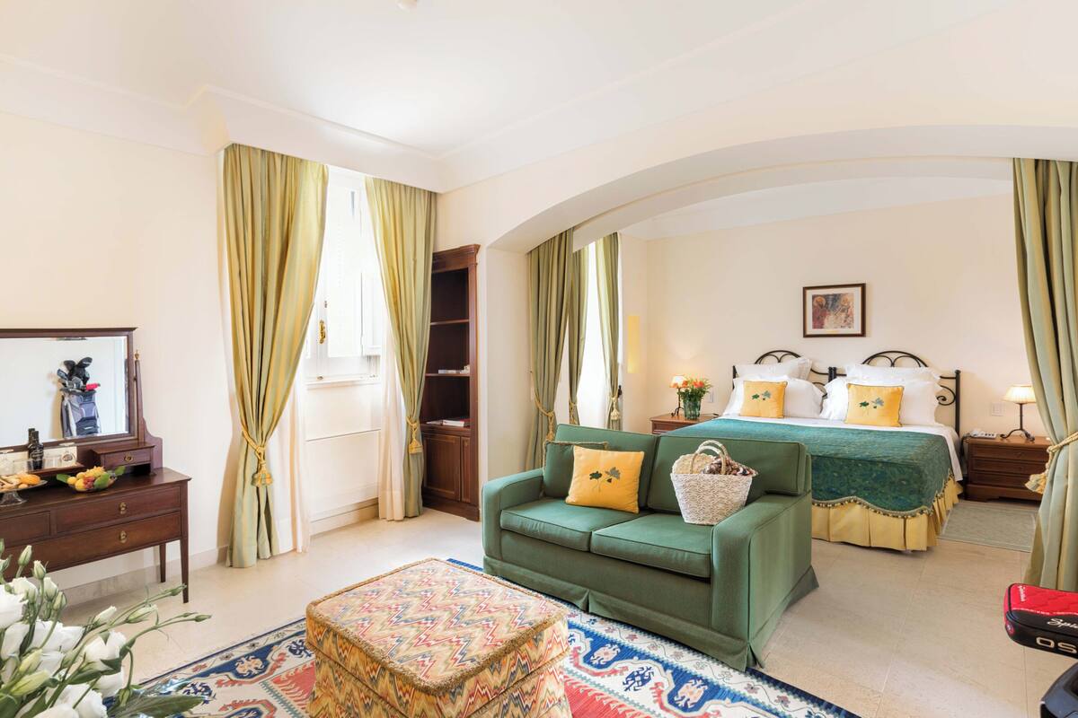 Una habitación con un sofá verde, una cama con cubrecama verde, un aparador de madera y un reposapiés estampado.