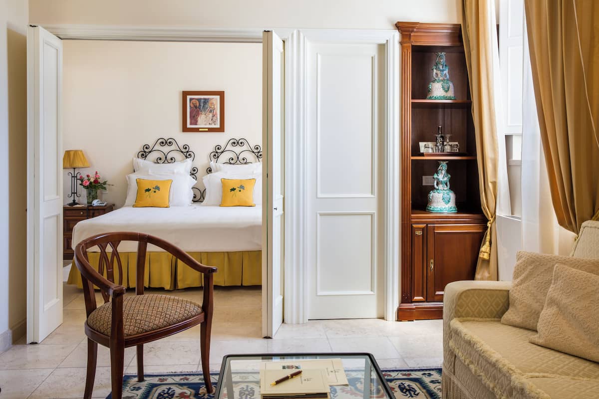 Habitación de hotel con cama, una silla de madera, sofá, mesa de centro de vidrio y estantería.