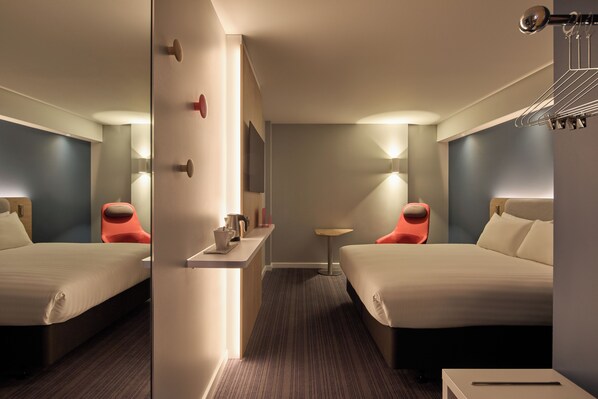 A hotel room with two beds, a small table, a chair, a mirror, and a wall-mounted shelf with a kettle and cups. - Holiday Inn Express London - Limehouse by IHG (London)
