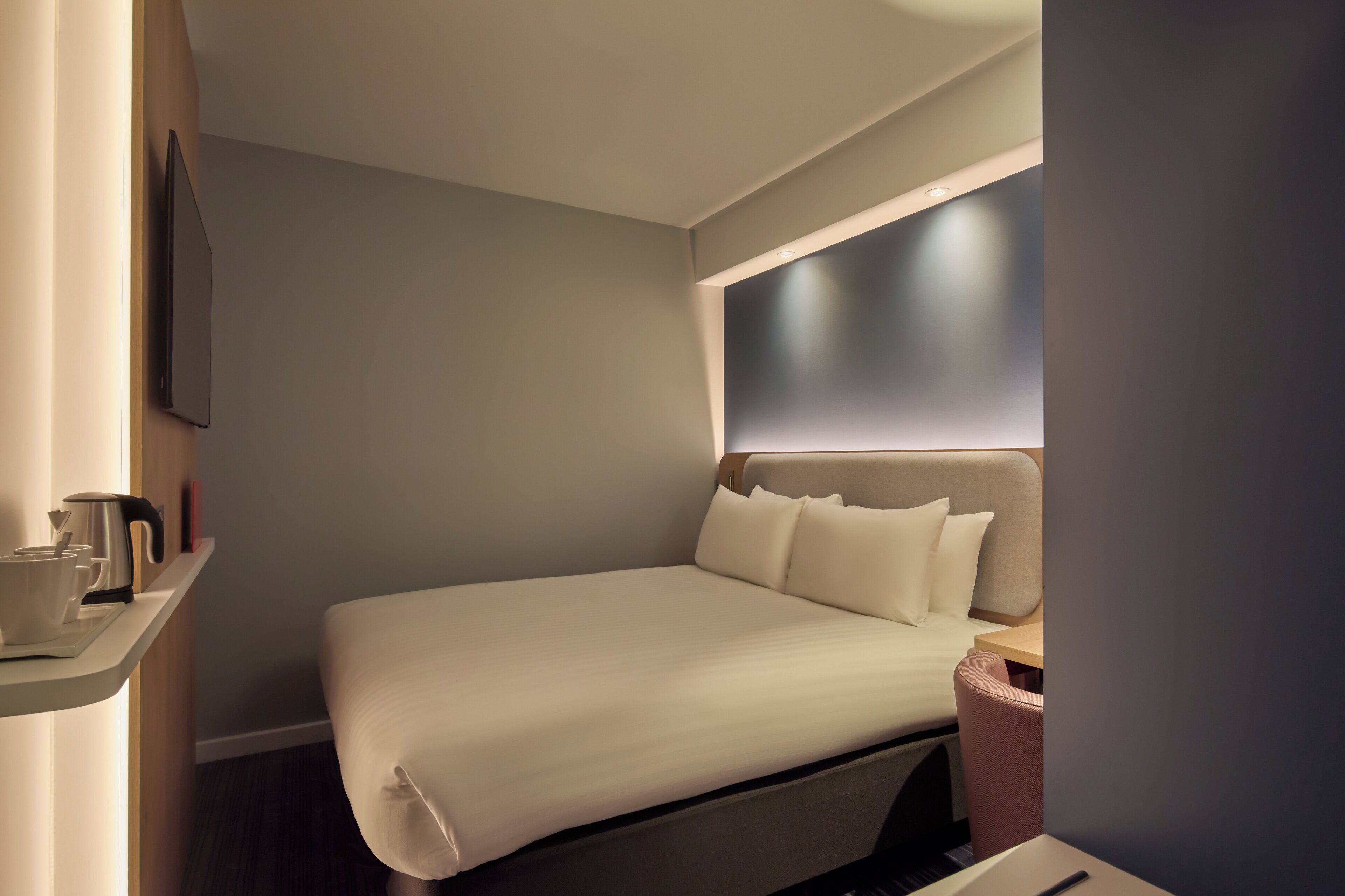 Habitación de hotel moderna con una cama bien hecha, una mesita de noche con hervidor y un televisor montado en la pared.