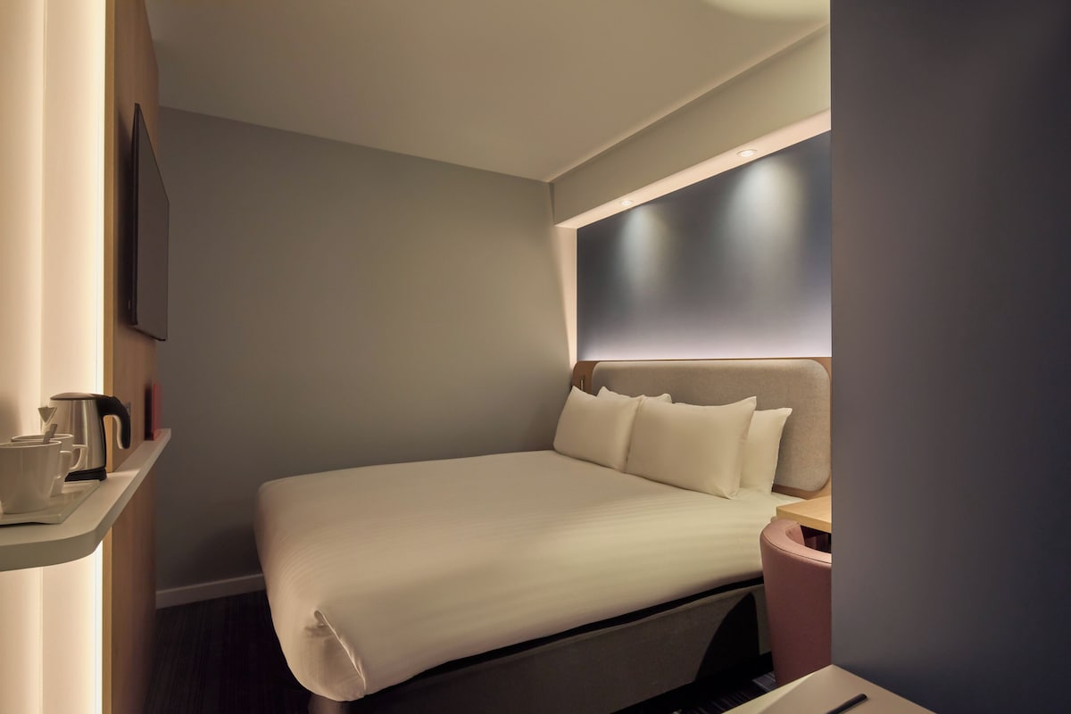 Habitación de hotel moderna con una cama bien hecha, una mesita de noche con hervidor y un televisor montado en la pared.