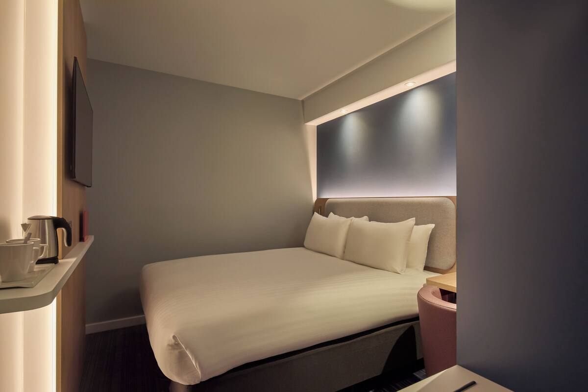 A modern hotel room with a neatly made bed, a bedside table with a kettle, and a wall-mounted TV.
