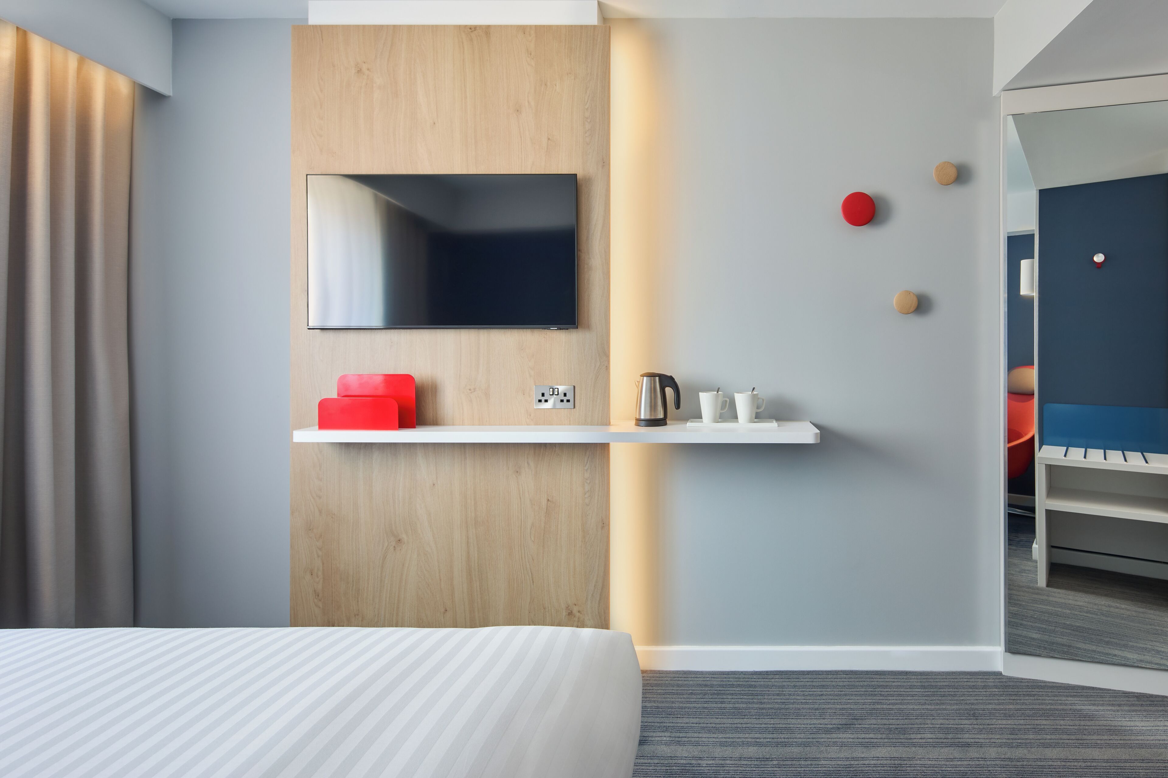 A modern hotel room with a large bed, a mounted TV, a wooden accent wall, a shelf with a kettle and cups, and a red object on the shelf.