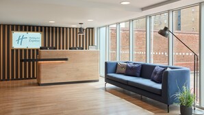 A modern hotel reception area with a wooden reception desk, a blue sofa, and large windows offering a view of a brick building. - Holiday Inn Express London - Limehouse by IHG (London)
