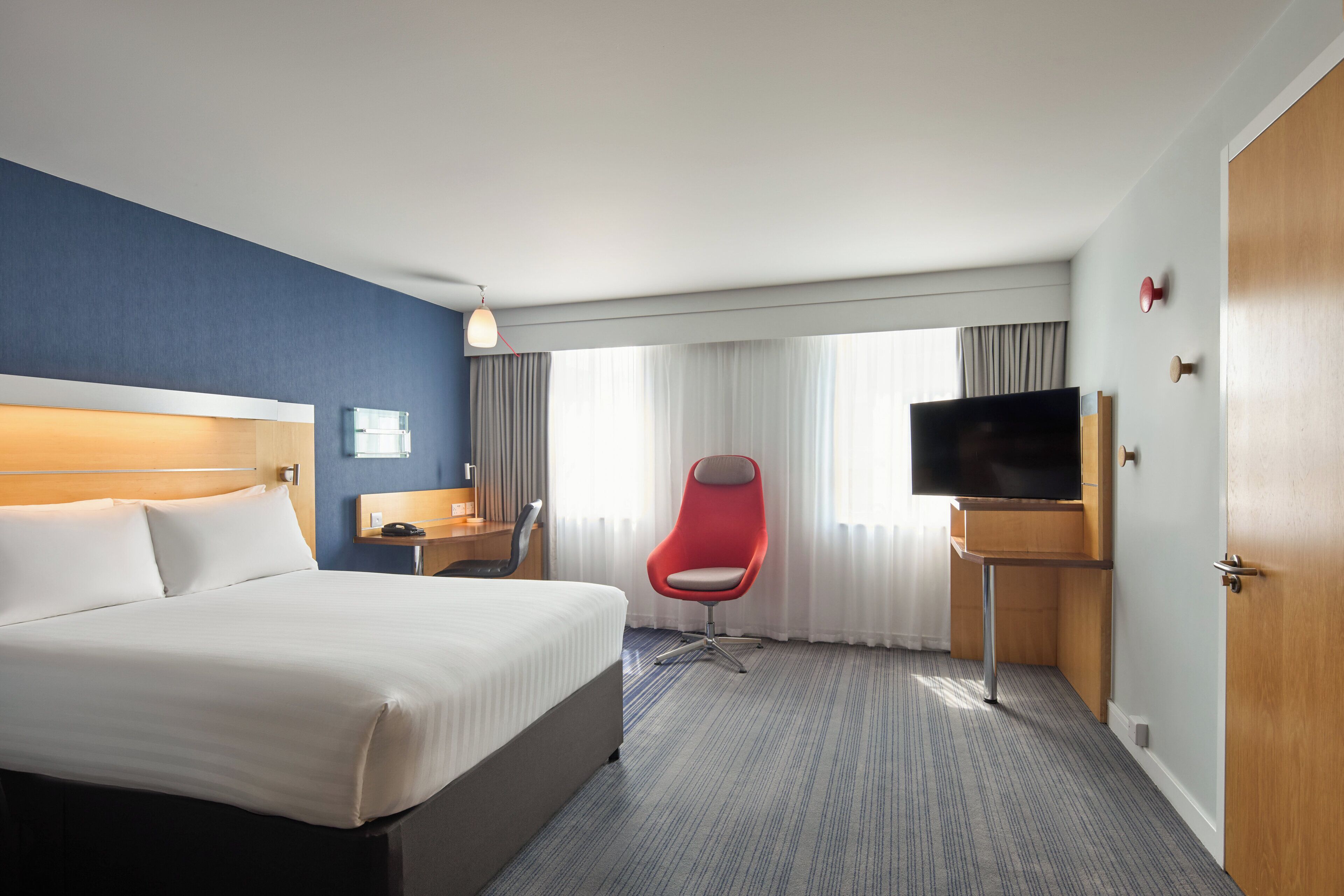 A hotel room with a large bed, a red chair, a TV, and a desk with a lamp.