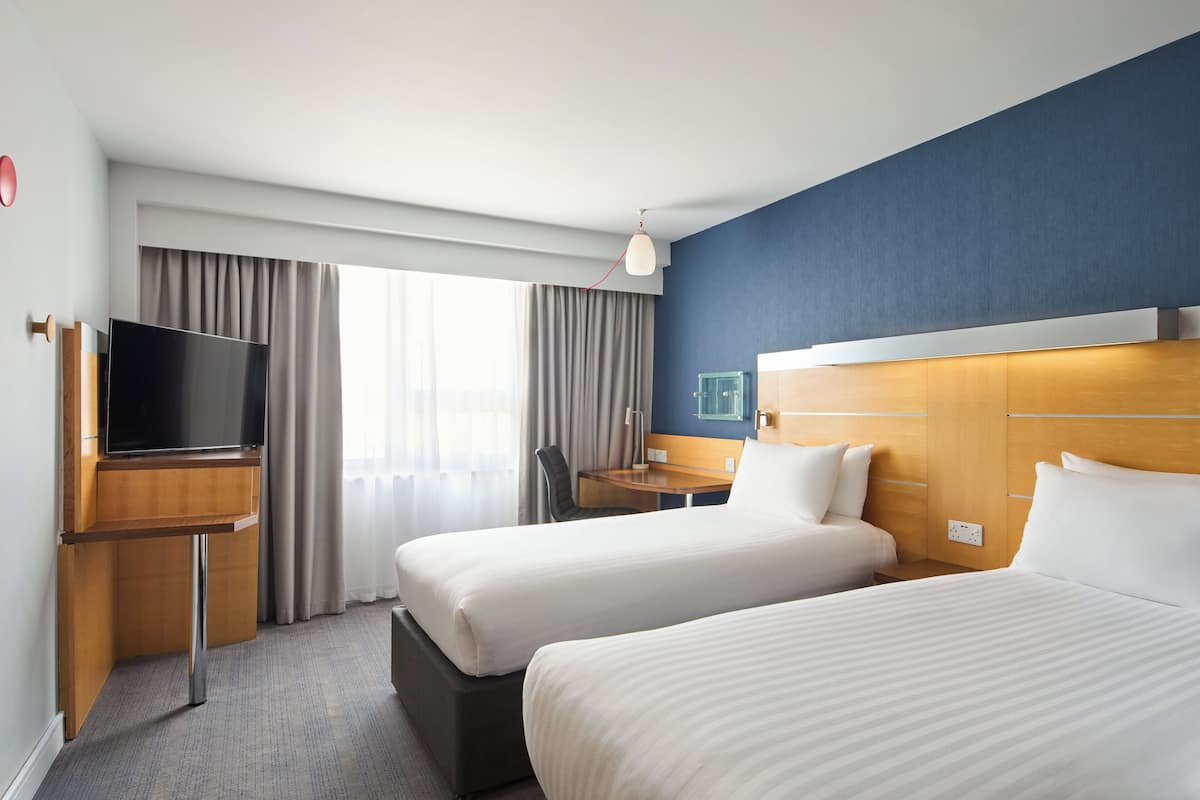 A hotel room with two beds, a desk with a TV, a window with curtains, and a wooden headboard.