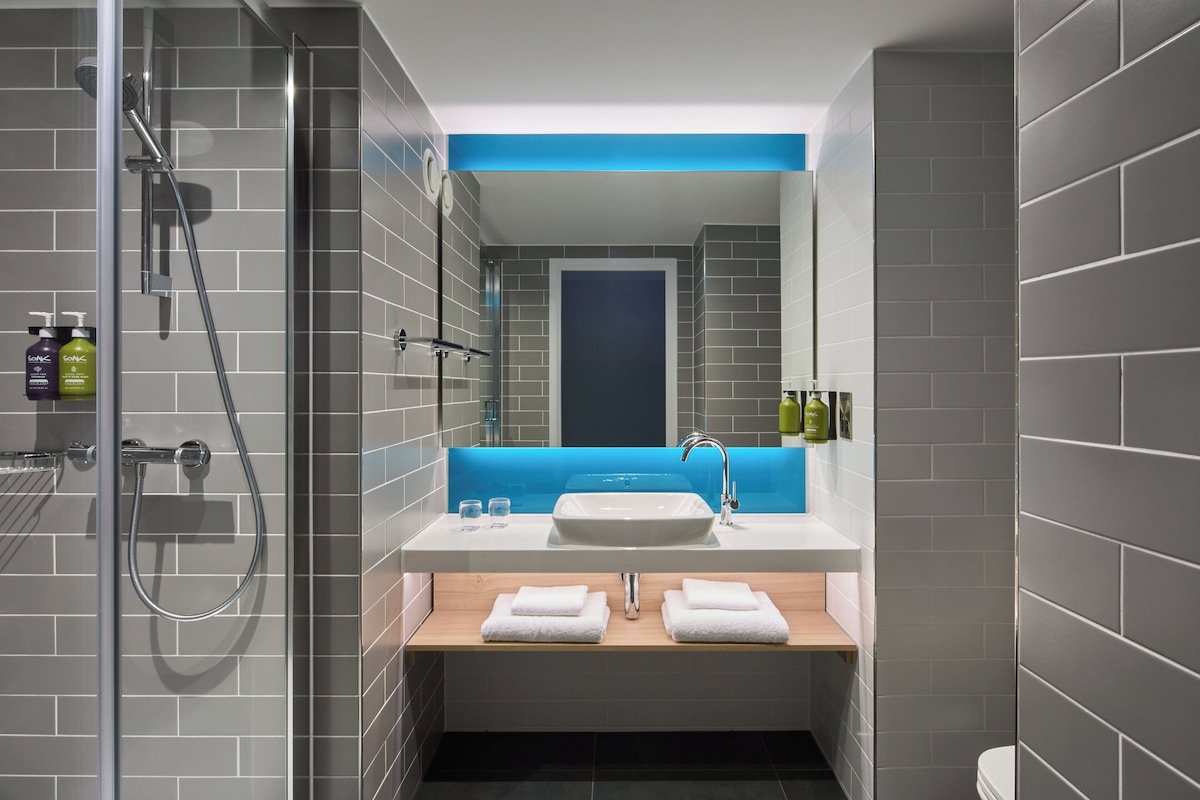 Un baño moderno con cabina de ducha de vidrio, un lavamanos blanco con detalles azules y toallas ordenadas sobre un estante de madera.