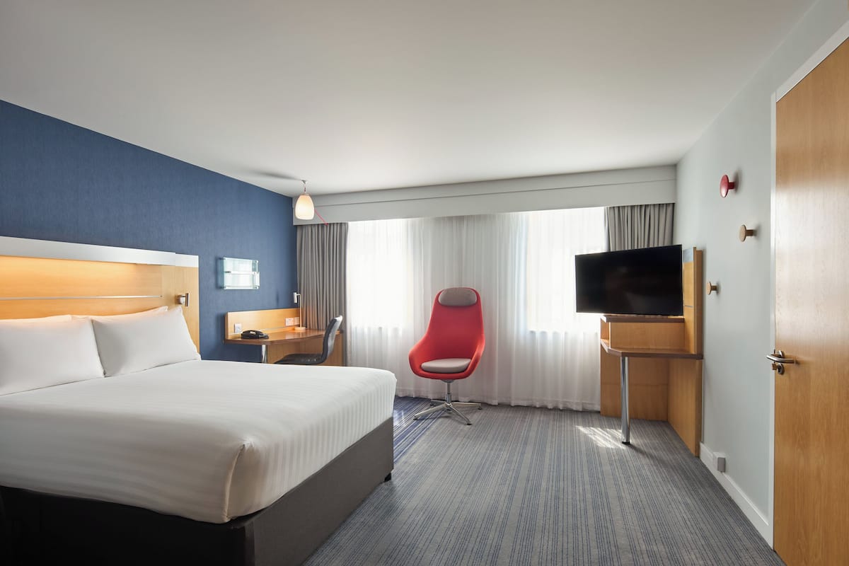 A hotel room with a large bed, a red chair, a TV, and a desk with a lamp.