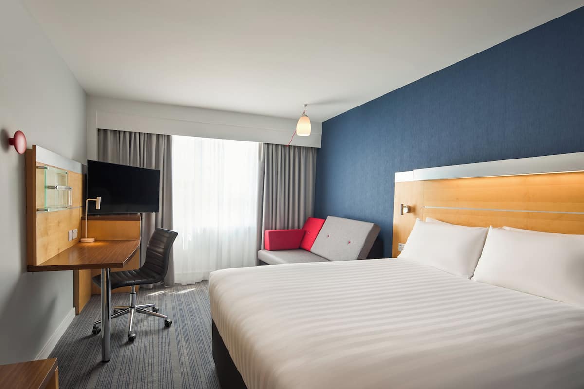 A hotel room with a bed, a desk with a TV, a chair, and a red sofa.